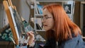 Inspired funny focused artist woman with red hair girl painter wears glasses holds painting brushes in mouth paints with Royalty Free Stock Photo