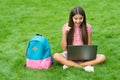 Inspired happy child using computer for online school study sitting in park on green grass with backpack, education Royalty Free Stock Photo