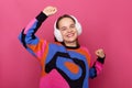 Inspired jocund girl wearing colorful jumper and fur earmuffs, dancing isolated on pink background, gorgeous young female model