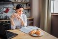 Intelligent adult woman sit at table in kitchen. She read journal and hold white cup with hot drink. Woman look down