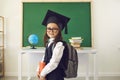 Intelligent little girl in graduation mortar hat posing at classroom. Smart child with backpack on first day of school