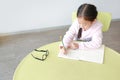 Intend little Asian girl writes in a book or notebook with pencil sitting on kid chair and table in classroom