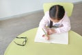 Intend little Asian girl writes in a book or notebook with pencil sitting on kid chair and table in classroom