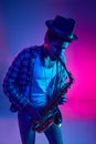 Intense musician, African-American man in hat playing saxophone in vivid lighting against pink-blue background. Royalty Free Stock Photo