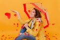 Interested brown-haired girl enjoying heart rain. Smiling attractive woman in yellow coat sitting on the floor with