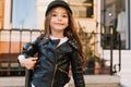 Interested slim little girl with long dark hair posing in front of school building next iron pillar. Portrait of