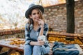 Interested young lady with cup of coffee posing in park in october day. Adorable woman in hat with black manicure