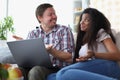 Interethnic family couple laughing and chatting while sitting on sofa at home with laptop. Royalty Free Stock Photo