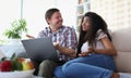 Interethnic family couple laughing and chatting while sitting on sofa at home with laptop. Royalty Free Stock Photo