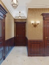Interior design of a classic hall corridor with yellow wallpaper, brown doors and wood paneling. Backlit paintings and sconces on