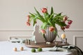 Interior design of easter dining room with colorful easter eggs, easter bunny sculptures, vase with tulips, wooden trace, beige