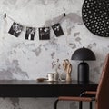 Interior design of industrial home office with black desk, brown leather armchair, pictures on a string, stylish lamp, gray