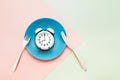 Intermittent fasting concept. White alarm clock on empty blue dish with knife and fork on pink-green background. top view, copy