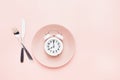 Intermittent fasting concept. White alarm clock on empty pink dish with knife and fork on pink background. top view, copy space Royalty Free Stock Photo