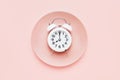 Intermittent fasting concept. White alarm clock on empty pink plate. Top view, copy space