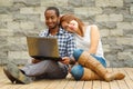 Interracial charming couple wearing casual clothes sitting down on wooden surface lookin at laptop together, in front of Royalty Free Stock Photo