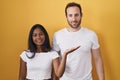 Interracial couple standing over yellow background smiling cheerful presenting and pointing with palm of hand looking at the Royalty Free Stock Photo