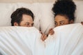 Interracial, happy and relaxed couple lying in bed, bonding and looking shy while hiding after waking up together in the