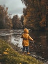 An intrepid young explorer clad in a yellow jacket ventures to fish along a leaf-strewn riverbank, embracing the crisp