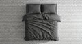 A neatly made bed with dark grey duvet and two matching pillows rests centrally on a textured white brick wall surface, offering a Royalty Free Stock Photo
