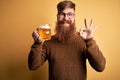 Irish redhead man with beard drinking a glass of refreshing beer over yellow background doing ok sign with fingers, excellent Royalty Free Stock Photo