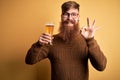 Irish redhead man with beard drinking a glass of refreshing beer over yellow background doing ok sign with fingers, excellent Royalty Free Stock Photo