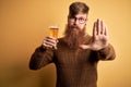 Irish redhead man with beard drinking a glass of refreshing beer over yellow background with open hand doing stop sign with Royalty Free Stock Photo