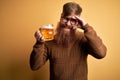 Irish redhead man with beard drinking a glass of refreshing beer over yellow background stressed with hand on head, shocked with Royalty Free Stock Photo