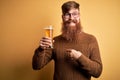 Irish redhead man with beard drinking a glass of refreshing beer over yellow background with surprise face pointing finger to Royalty Free Stock Photo