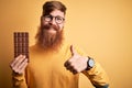 Irish redhead man with beard eating a sweet chocolate bar over yellow isolated background happy with big smile doing ok sign, Royalty Free Stock Photo