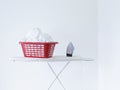 Iron and laundry basket on ironing board against white wall Royalty Free Stock Photo