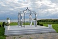 Iron pavilion decorated with white cloths for a wedding ceremony on a platform on the lake shore under a cloudy sky, copy space