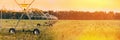 Irrigation Machine At Agricultural Field With Young Sprouts, Green Plants On Black Soil. Panoramic View Of Sunset Sky