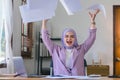 Irritated Beautiful muslim millennial businesswoman wearing a hijab sitting at office desk throwing up papers with