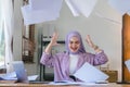 Irritated Beautiful muslim millennial businesswoman wearing a hijab sitting at office desk throwing up papers with