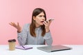 Irritated frustrated business woman in striped shirt leaving voice message holding phone near mouth sitting at workplace, giving Royalty Free Stock Photo