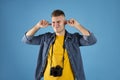 Irritated male traveler with photo camera covering his ears with fingers on blue studio background