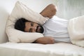 Irritated young indian man lying in bed and covering ears with pillow Royalty Free Stock Photo