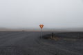 Iceland yield sign at junction of volcanic gravel road with tarmac road with some wire fence on foggy day