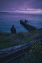 An isolated man sits on the grassy hillside overlooking an old broken pier Royalty Free Stock Photo