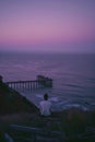 An isolated man sits on the grassy hillside overlooking an old broken pier Royalty Free Stock Photo