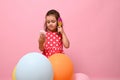 Isolated portrait on pink background of a gorgeous baby girl in stylish pink dress, standing behind multicolored balloons, combing