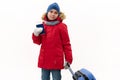 Happy traveler boy in red parka, jeans, wool mittens, goes for winter holidays, walks with air ticket and blue suitcase Royalty Free Stock Photo
