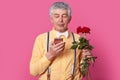 Isolated shot of attractive old man focused in smartphone, recives message from lover, agree to have date, carries red roses,