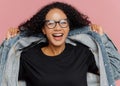  shot of curly cheerful woman tries on new denim jacket, wears optical glasses, smiles broadly, has perfect mood, poses