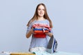 Isolated shot of pretty female with long hair, keeps pile with laundry, going to iron with electric iron, isolated over blue backg