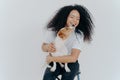 Isolated shot of smiling Afro American woman with curly hairstyle, gets kiss from favourite dog, sits at chair against white wall Royalty Free Stock Photo