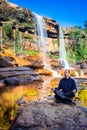 Isolated young man meditating near waterfall falling from mountain top with reflection and blue sky Royalty Free Stock Photo