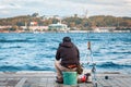Man with equipment sitting on a bucket on coast and fishing while talking on phone