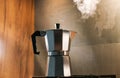 An Italian espresso mokka machine with steam getting out of it while making a fresh coffee in a regular kitchen with wood and ston